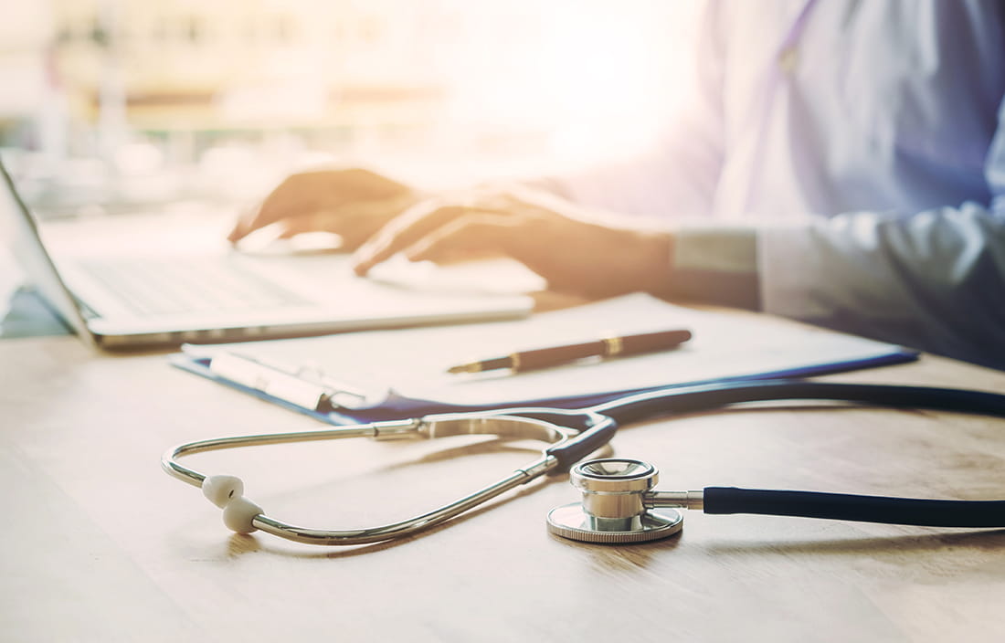 Up close shot of a person working on their laptop with a stethoscope laying next to it.