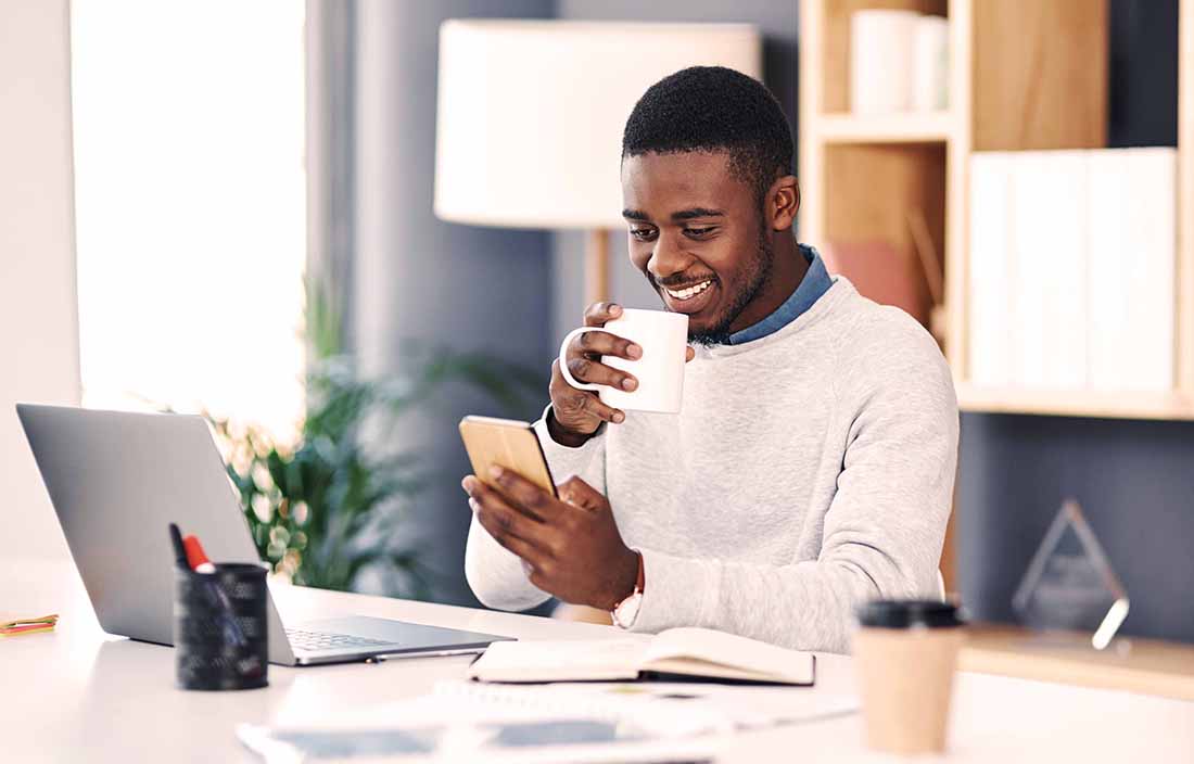 Man sitting at desk looking at his phone and drinking coffee