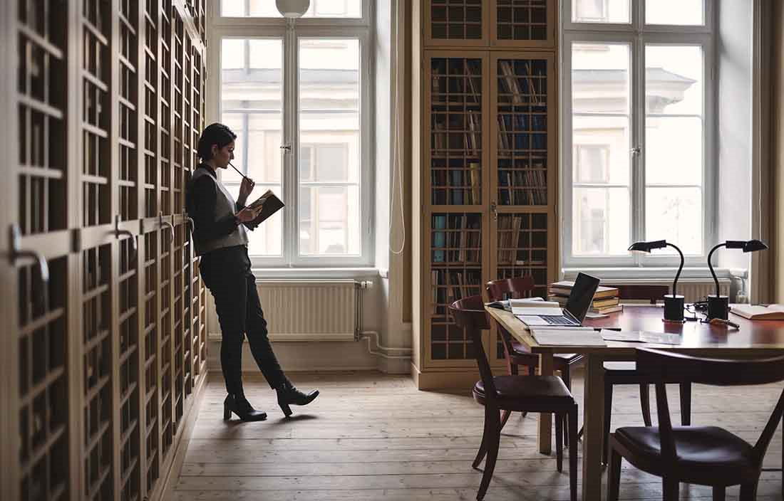 Woman leaning against a bookshelf reading a book.