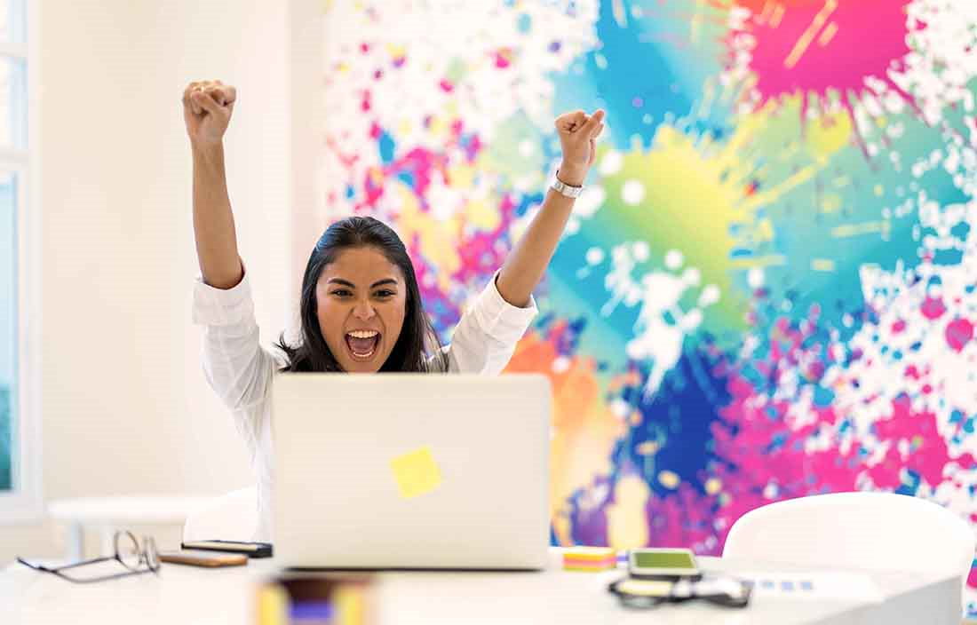 Woman sitting in front of laptop with her hands in the air celebrating.
