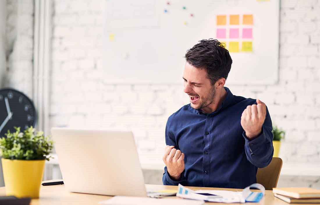 Businessman working from his home office celebrating at his kitchen table with a laptop in front of him.