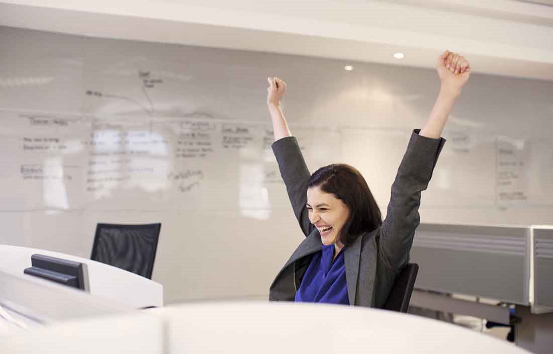 Businessperson celebrating in the office at their desk with their arms up in the air.