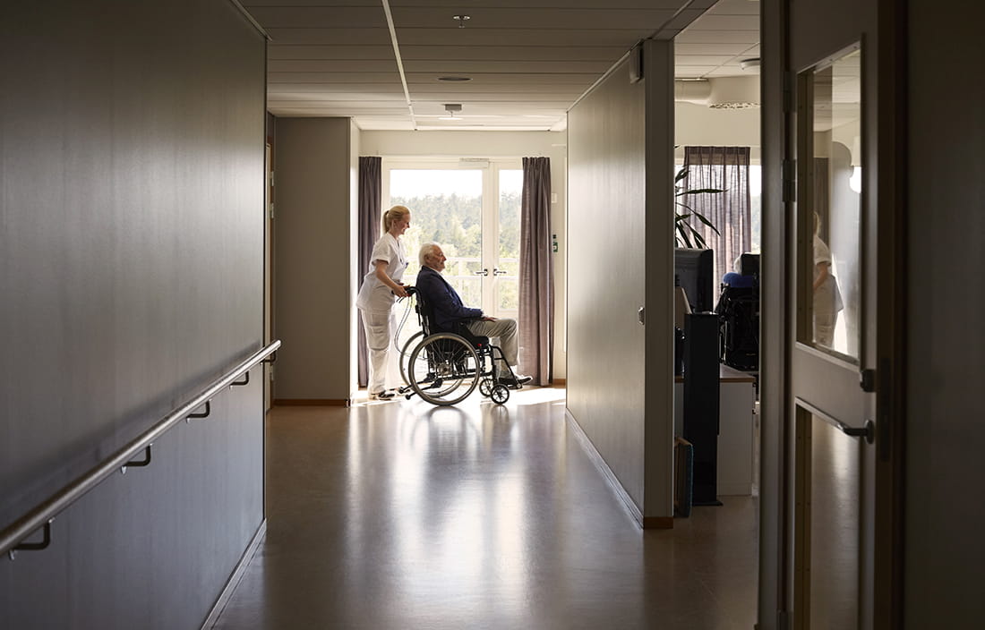 Nurse pushing an elderly man in a wheelchair through a hallway.