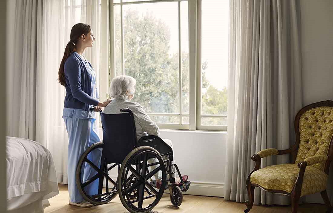 Healthcare worker standing behind wheelchair for elderly patient.