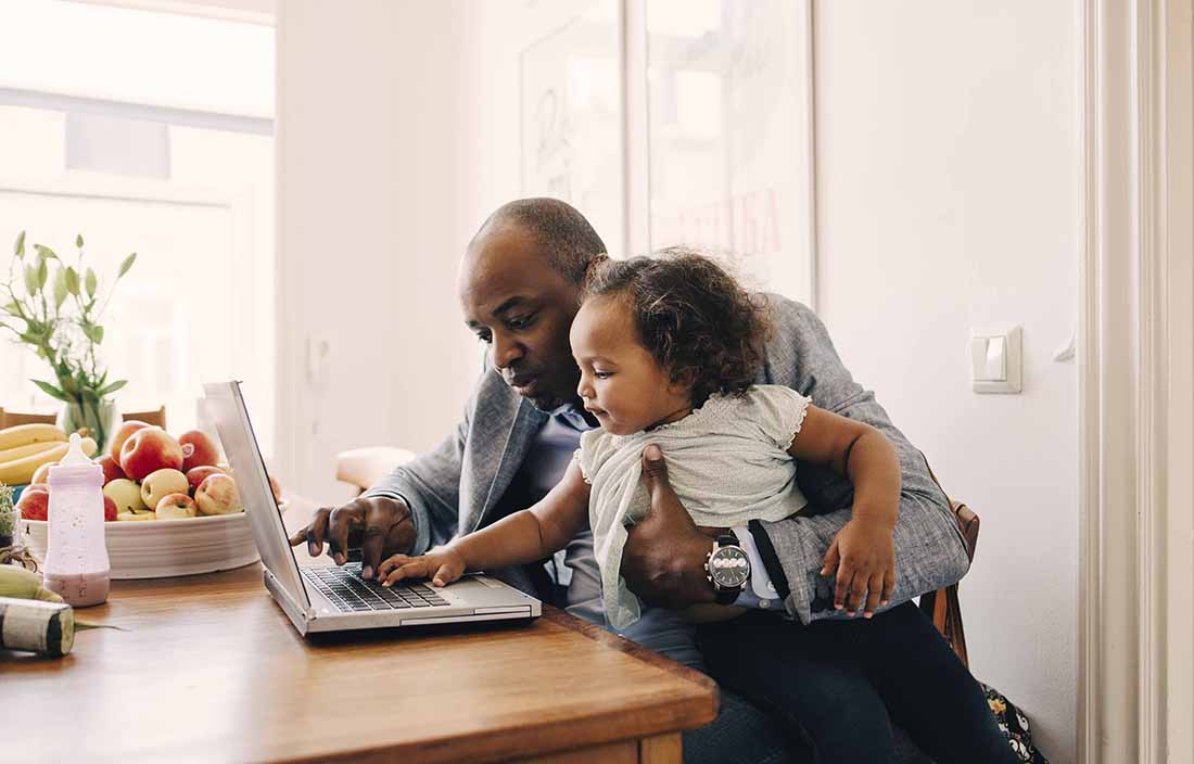 Dad holding his young toddler while using a laptop computer.