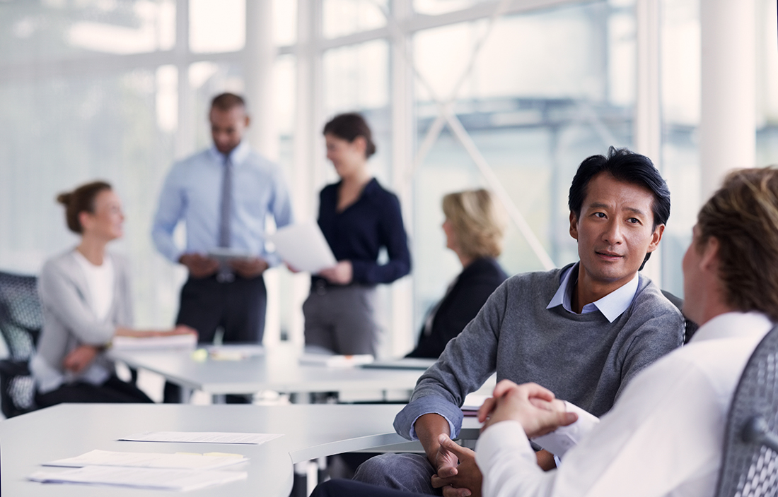 Picture of a meeting room with two people talking in the foreground with a group of people talking in the background.