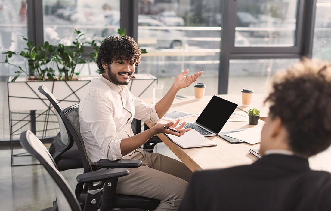 Smiling businessman talking to blurred colleague near laptop and notebook in a modern, open office