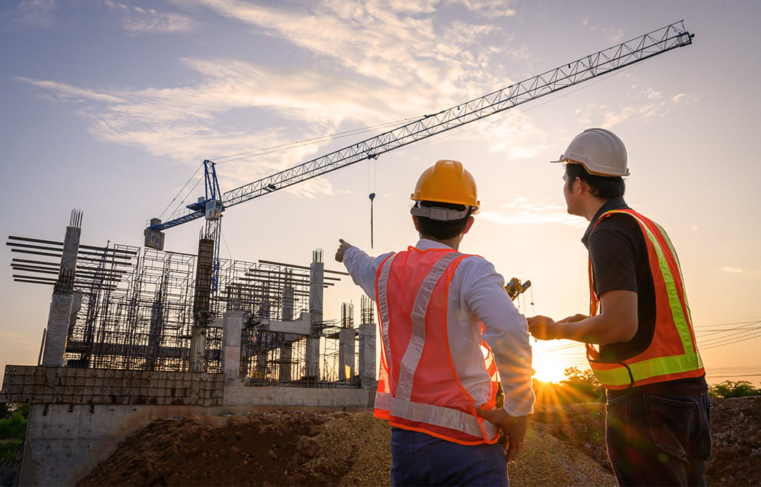 Two construction workers in the foreground, one pointing to an industrial building under construction, at either dawn or dusk