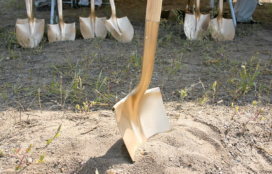 Foreground with golden shovel sticking up from mount of dirt, with background row of shovels and people's feet.