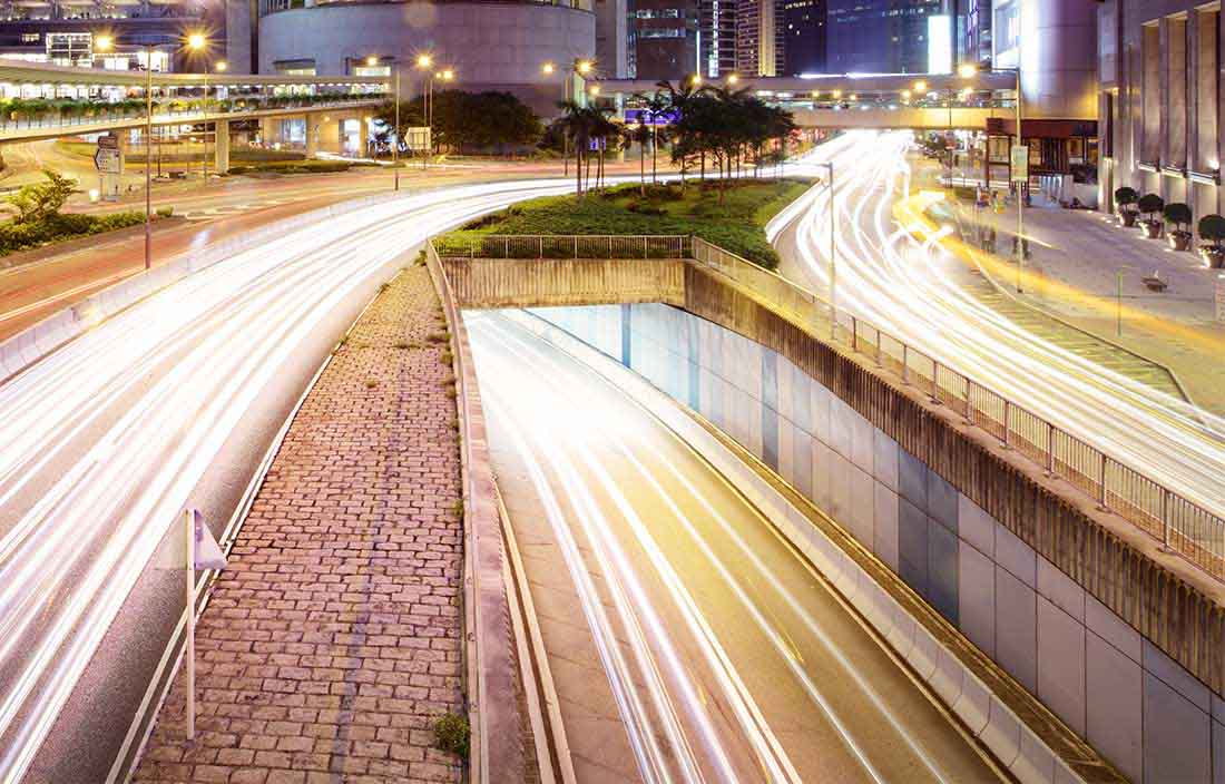 Time lapse image of vehicles driving under a road bridge at night next to industrial and office buildings