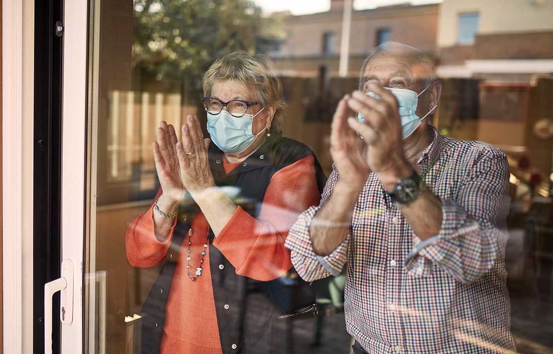 Two seniors with COVID medical masks looking out their senior living home window and clapping