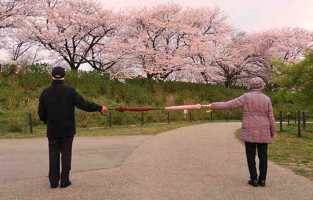 Two seniors walking down a senior living community lane holding out 2 umbrellas to stay 6 feet apart because if COVID