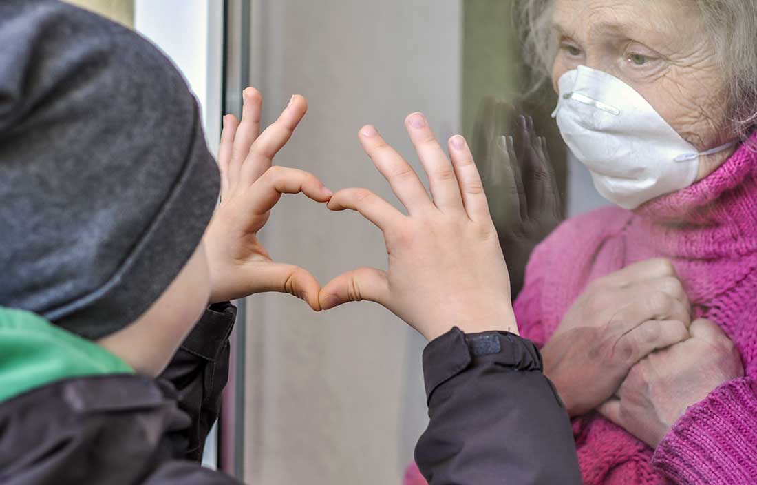 Senior living resident visits with her grandson through a window as he hold up his hands in a heart