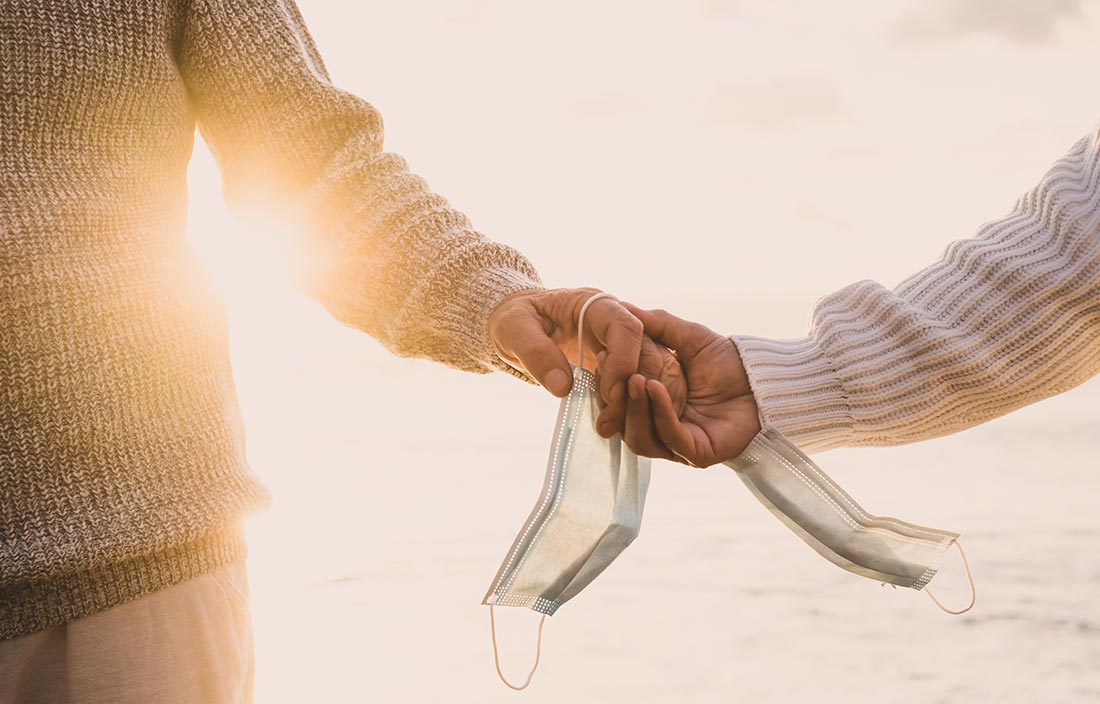 Close up photo of two seniors in sweaters holding hands with medical masks around their wrists. Sun glare is shining from around the figures, and the background is a body of water.