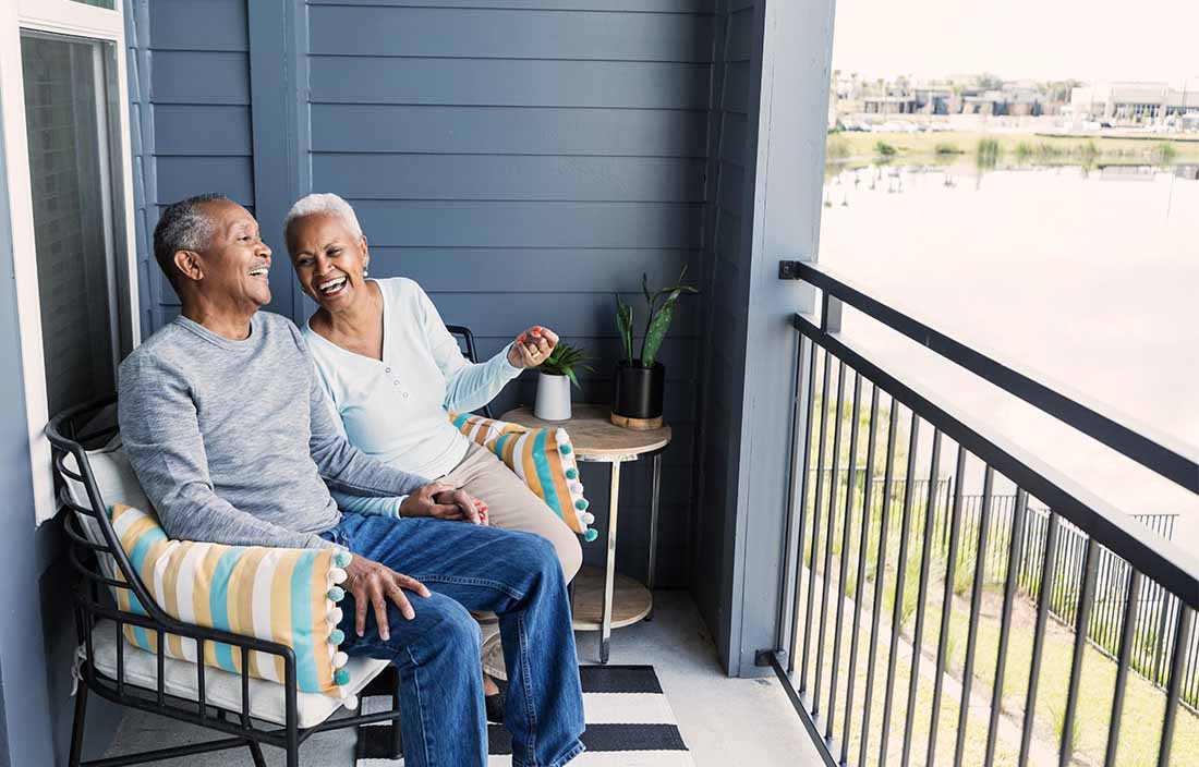 A senior African-American couple sitting side by side on their porch or balcony, relaxing, and holding hands, conversing and laughing together.