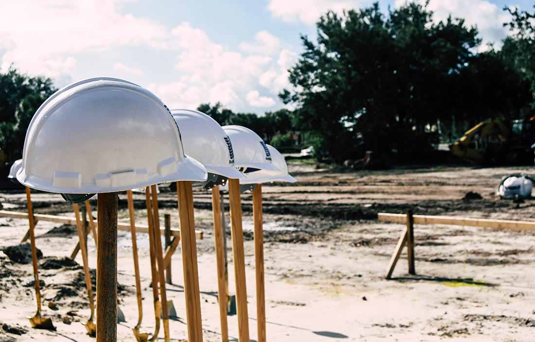 In the foreground, there are hardhats on shovels that are stuck in the ground and an open dirt field is ready for construction of a senior living construction project