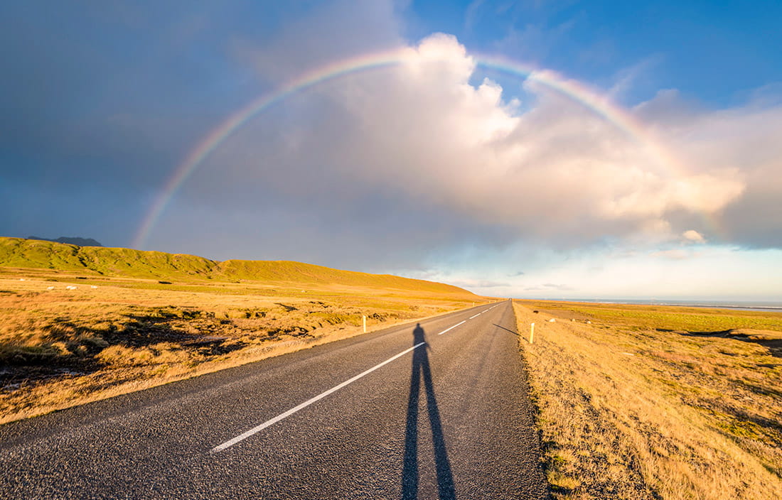 Shadow stretched out on a desert road leading to the horizon under a rainbow