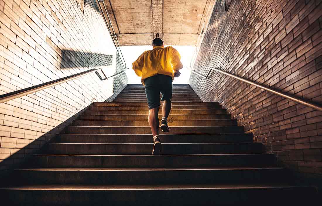 Senior man in a yellow jacket running up enclosed stairs toward a bright light