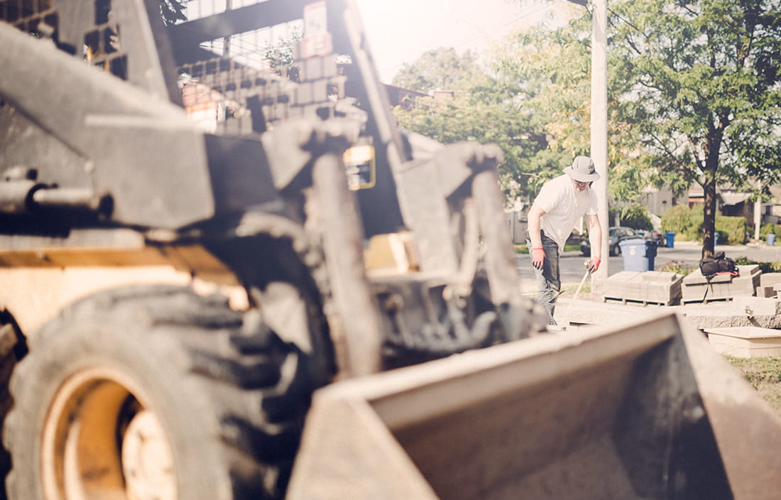 Bob cat construction machnery on the front yard of a senior living house. There is a construction worker in the back renovating the driveway pavement