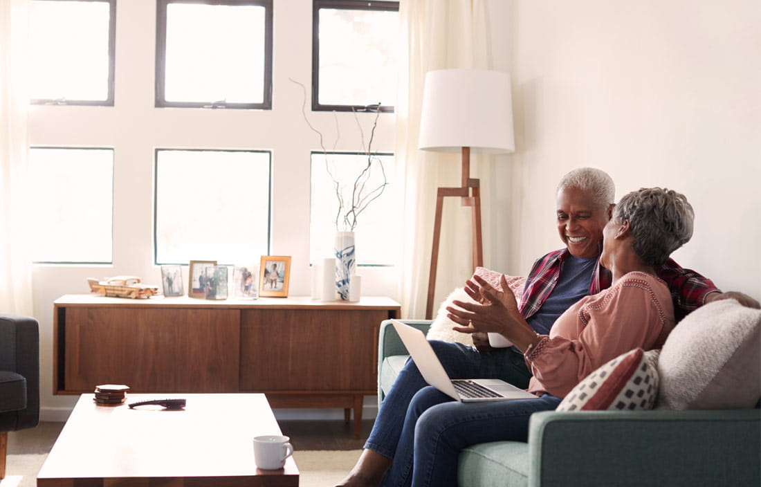 Senior couple on a living room couch with a laptop, laughing and gesturing as they research senior living communities online