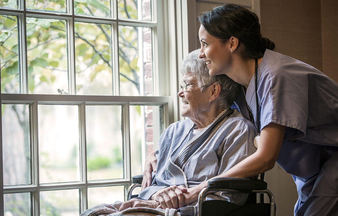 Caregiver and senior in a wheelchair smiling while looking out a window at a skilled nursing facility