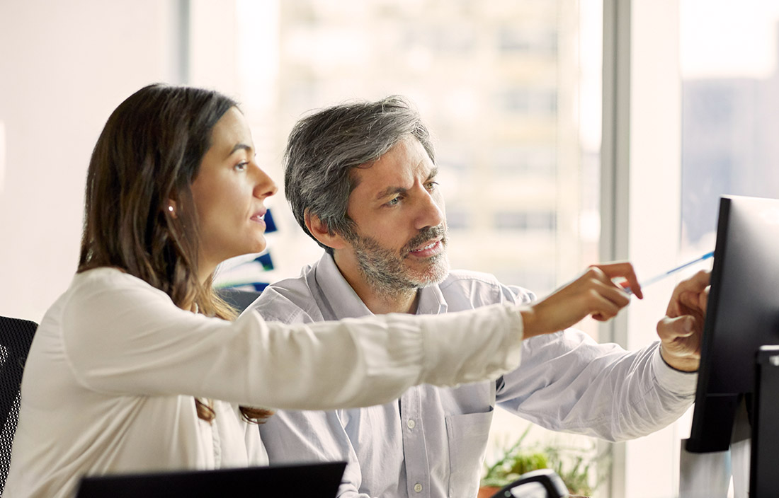 Colleagues, on woman and one man, pointing to a computer on an office desk where they're sitting and discussing senior living market trends and data for operations and repositioning decisions