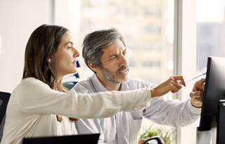 Colleagues, on woman and one man, pointing to a computer on an office desk where they're sitting and discussing senior living market trends and data for operations and repositioning decisions