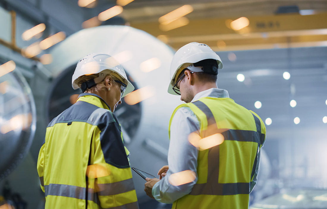 Two Heavy Industry Engineers Stand in Pipe Manufacturing Factory, Use Digital Tablet Computer, Have Discussion. Construction of Oil, Gas and Fuels Transport Pipeline. Back View Sparks Flying