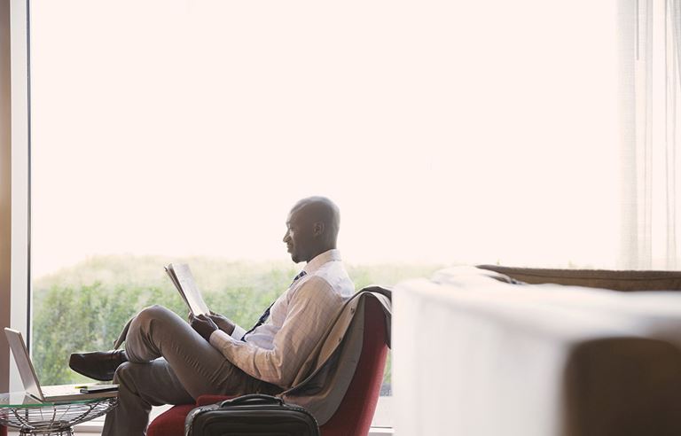 Image of a man sitting on a chair and reading a newspaper, with a window in the background.