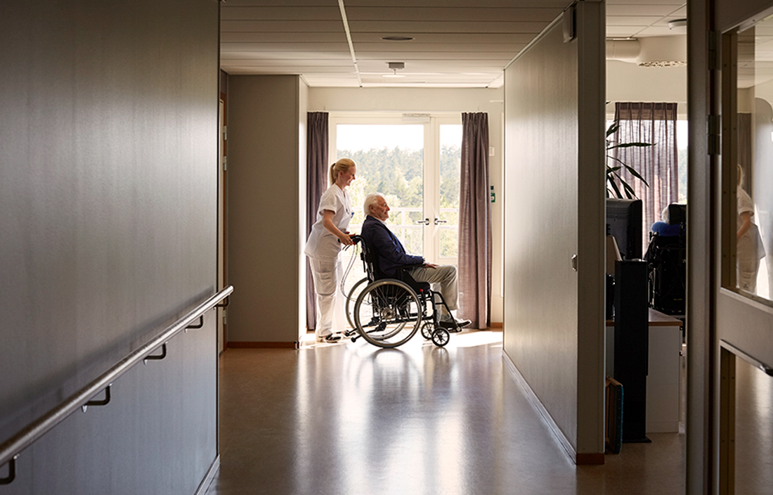Nursing assistant pushing an elderly senior citizen in a wheelchair through a hospital hallway.
