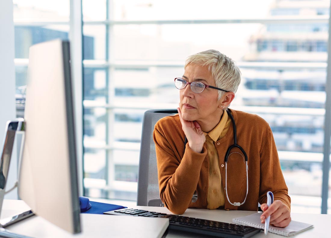 Elderly woman physician sitting at her desk using a desktop computer.