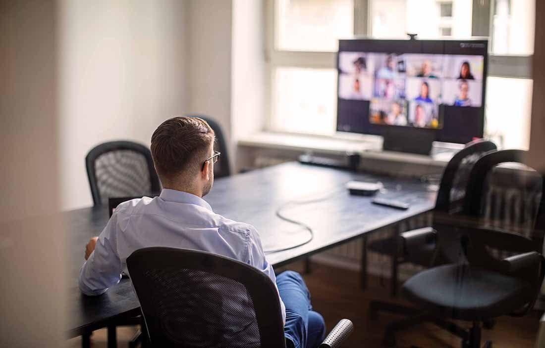 Businessman sitting at a conference table looking at a TV monitor with multiple members on a conference call.