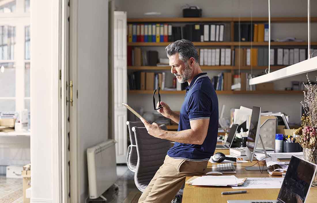 Man in casual business clothes leaning against a desk using a handheld tablet device.