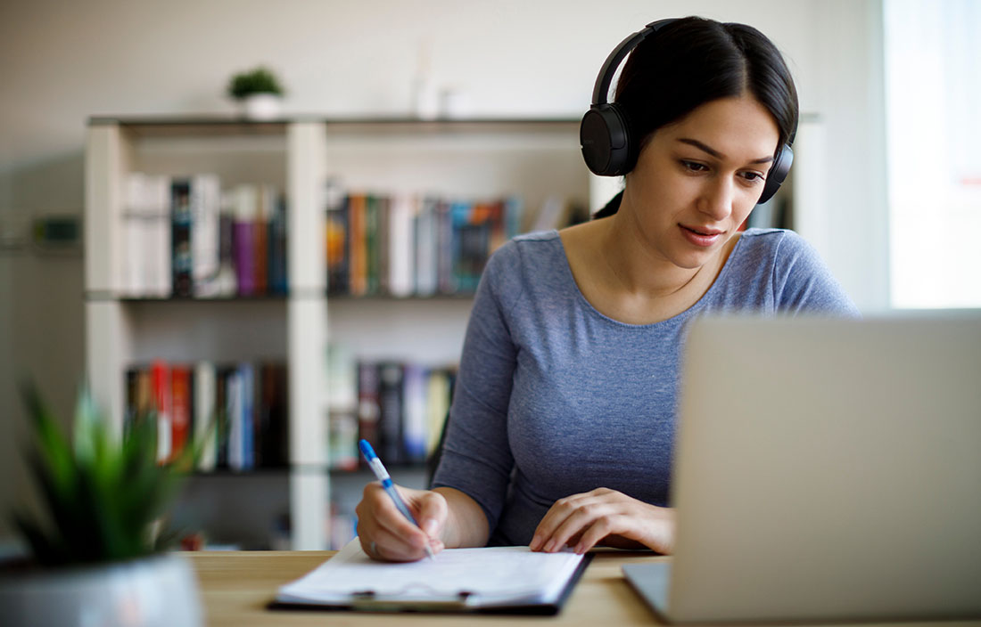 Businesswoman wearing headphones while taking notes in front of her laptop computer.