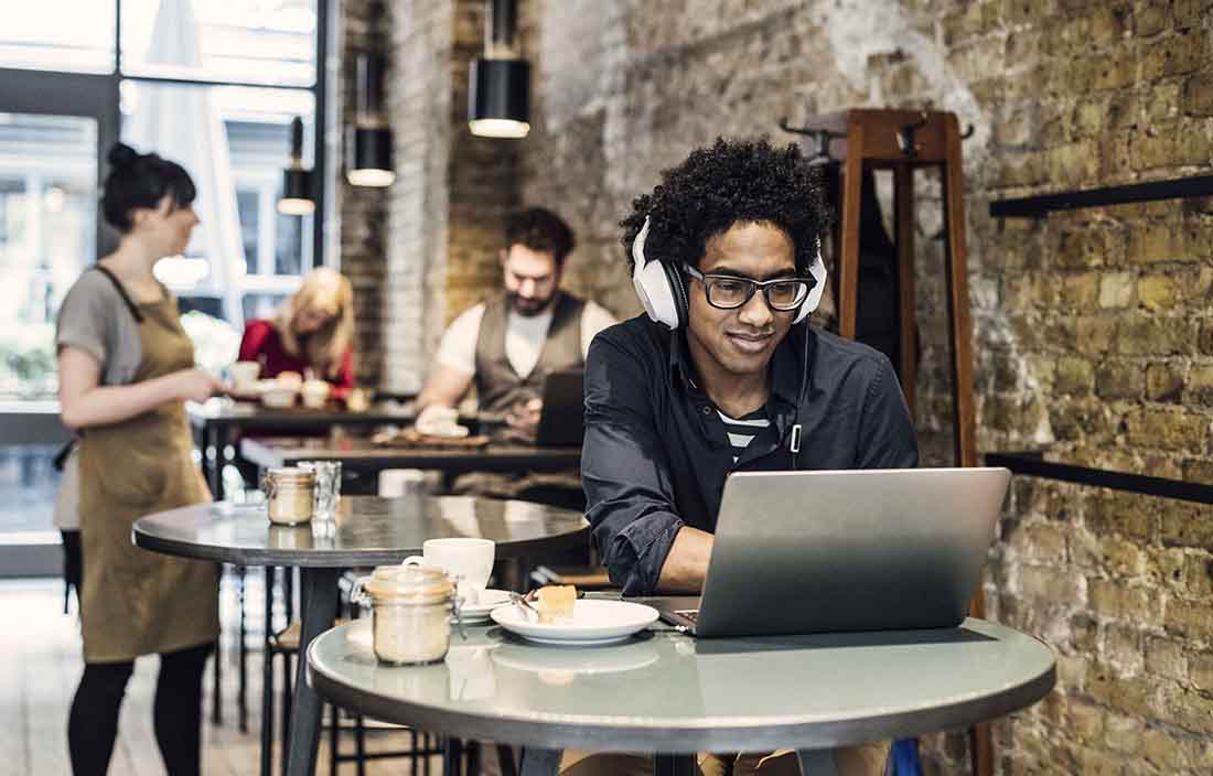 Business professional in casual clothes in a coffee shop wearing headphones while using a laptop computer.